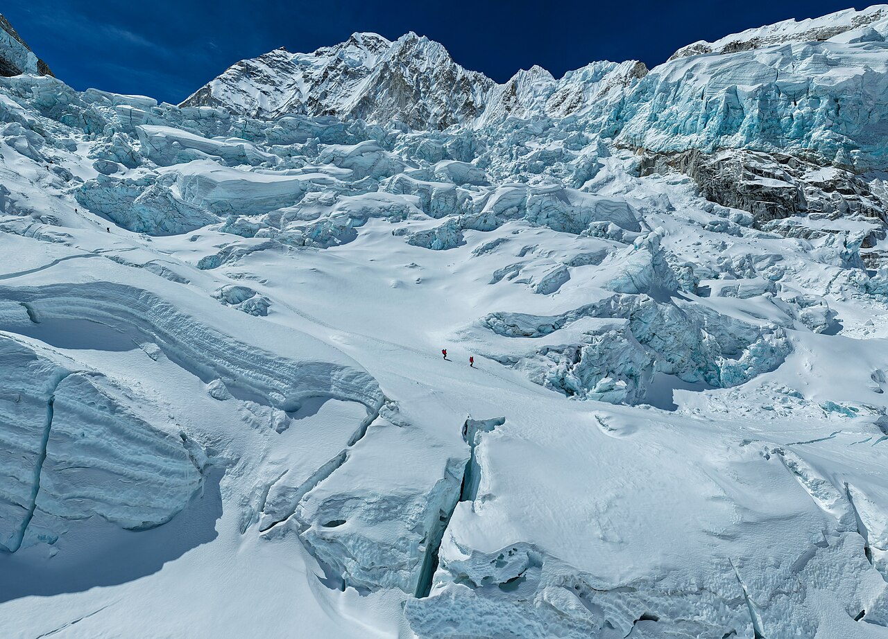 Climbers ascending a snowy high-altitude mountain pass with prayer flags and visible breath